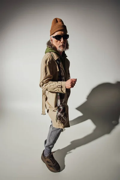 Старение со стилем, старший хипстер мужчина в темных солнцезащитных очках, шапка шапочка, зеленая толстовка и бежевый плащ глядя на камеру на сером фоне с тенью, положительное старение и концепция моды — стоковое фото