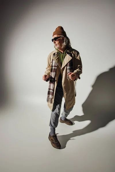 Personalidade expressiva, homem hipster sênior em óculos escuros, chapéu de gorro e casaco de trincheira bege em pé em pose elegante no fundo cinza com sombra, conceito de envelhecimento positivo e na moda — Fotografia de Stock