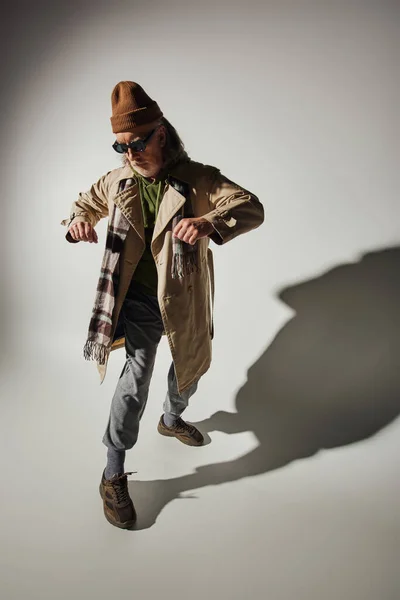 Старіння з концепцією стилю, повна довжина старшого хіпстерського чоловіка в темних сонцезахисних окулярах, капелюх бежевого траншеї пальто і плетений шарф, що стоїть в стильній позі на сірому фоні з тіні — стокове фото