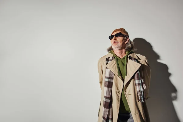 Пожилой человек в стильной повседневной одежде и темных солнцезащитных очках стоя на сером фоне с тенью и глядя в сторону, хипстерский тренд, шапочка шапочка, бежевый плащ, мода и возрастная концепция — стоковое фото
