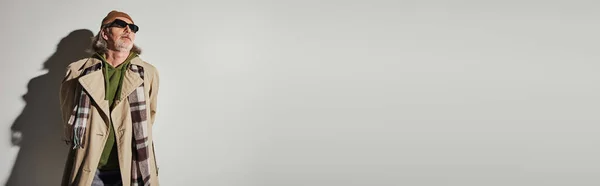 Homme âgé en vêtements décontractés tendance et lunettes de soleil sombres debout sur fond gris avec ombre et détournement des yeux, style hipster, bonnet, trench coat beige, écharpe à carreaux, bannière avec espace de copie — Photo de stock