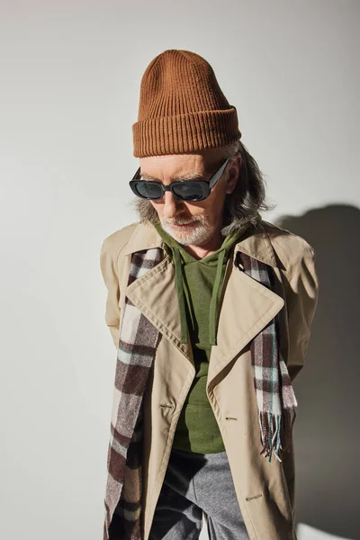 Senior male model, hipster style clothes, beanie hat, dark sunglasses, plaid scarf, beige trench coat, individuality, fashion and age concept, grey background with shadow — Stock Photo