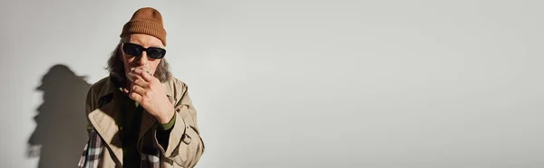 Modelo masculino estilo hipster sênior em óculos escuros, chapéu beanie, casaco de trincheira bege e cachecol xadrez segurando a mão perto do rosto e olhando para a câmera no fundo cinza com sombra, banner com espaço de cópia — Fotografia de Stock