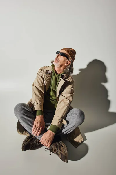 Homme hipster senior en bonnet, lunettes de soleil sombres et trench coat beige assis avec jambes croisées et regardant loin sur fond gris avec ombre, concept de vieillissement à la mode — Photo de stock