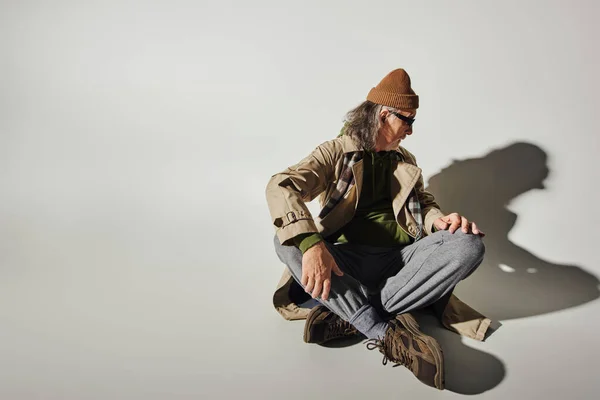 Mann im trendigen Hipster-Stil mit Hut, dunkler Sonnenbrille, beigem Trenchcoat und Turnschuhen, der seinen eigenen Schatten betrachtet, während er mit überkreuzten Beinen auf grauem Hintergrund mit Kopierraum sitzt — Stockfoto