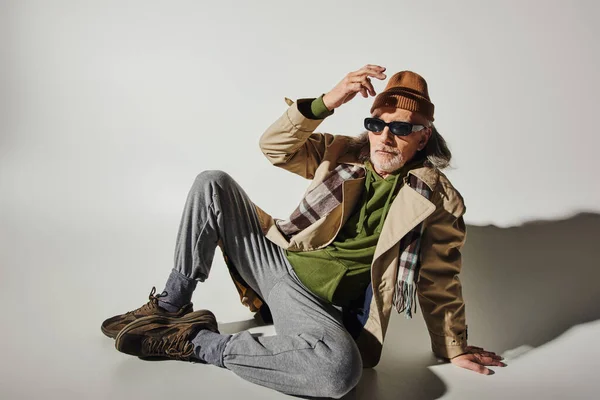 Full length of expressive and trendy senior man in beanie hat, dark sunglasses, beige trench coat and sneakers sitting on grey background with shadow, hipster fashion, stylish lifestyle — Stock Photo