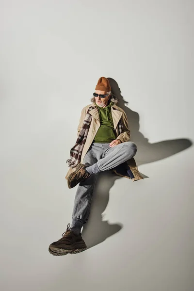 High angle view of senior male model in trendy hipster style attire and dark sunglasses laying and looking away on grey background with shadow, beanie hat, beige trench coat, fashion and age concept — Stock Photo
