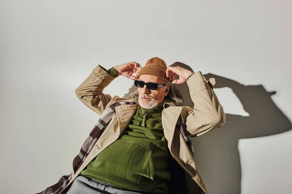 Aged man in dark sunglasses, green hoodie and beige trench coat adjusting beanie hat and looking away on grey background with shadow, fashionable and positive aging concept — Stock Photo