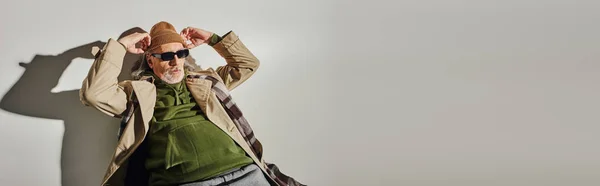 Hombre de estilo hipster de moda en gafas de sol oscuras y gabardina beige ajustando sombrero de gorro mientras posa sobre fondo gris con sombra, envejecimiento con concepto de estilo, bandera - foto de stock