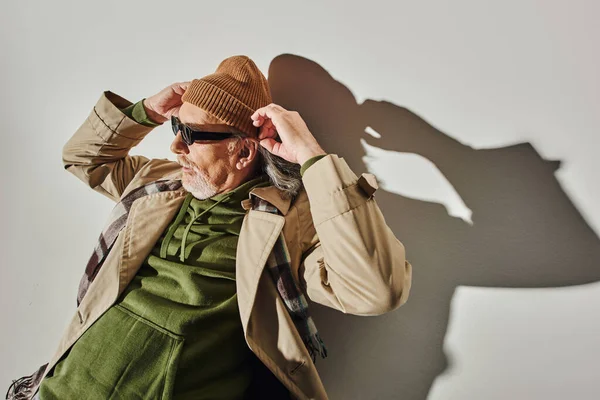 Vista de ángulo alto del hombre barbudo senior en gafas de sol oscuras y sudadera con capucha verde ajustando el sombrero de gorro y mirando hacia otro lado mientras está acostado sobre un fondo gris con sombra, moda hipster, concepto de envejecimiento positivo - foto de stock
