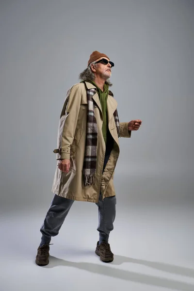 Full length of aged and trendy man in beanie hat, dark sunglasses, beige trench coat and plaid scarf looking away while standing on grey background, stylish pose, fashion shoot — Stock Photo