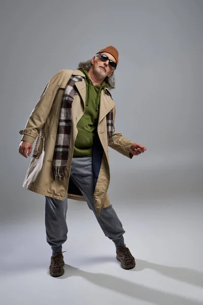 Full length of aged senior model in hipster style outfit posing on grey background, beanie hat, dark sunglasses, beige trench coat, sneakers, fashionable lifestyle and positive aging concept — Stock Photo