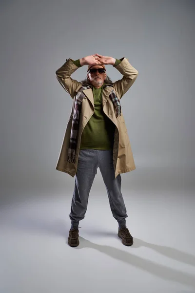 Full length of cool and senior hipster man in dark sunglasses, beanie hat, beige trench coat and sneakers posing with hands above head on grey background, fashionable aging concept — Stock Photo