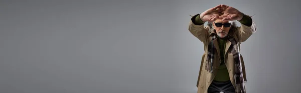 Homem de estilo moderno hipster em óculos escuros e casaco de trincheira bege posando com as mãos estendidas apertadas no fundo cinza, envelhecimento elegante, tiro de moda, banner com espaço de cópia — Fotografia de Stock