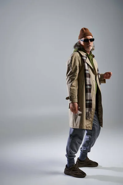 Full length of senior and trendy man in dark sunglasses looking at camera on grey background, beanie hat, beige trench coat, plaid scarf, sneakers, fashion and age concept — Stock Photo