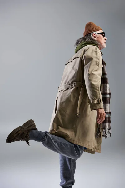 Side view of senior male model in beanie hat, dark sunglasses and beige trench coat posing on grey background, stylish pose, hipster fashion, trendy aging concept, fashion shoot — Stock Photo
