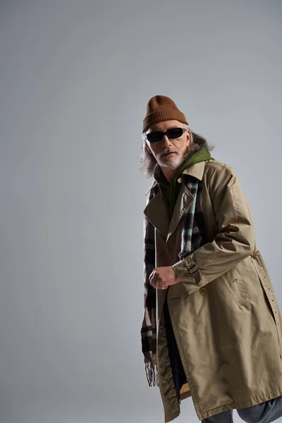 Сірошерстий старший чоловік в темних сонцезахисних окулярах, капелюх, бежева траншея пальто і плетений шарф, дивлячись на сірий фон, виразна особистість, хіпстерська мода, позитивна концепція старіння — стокове фото