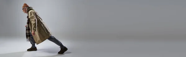 Vista lateral do homem idoso na moda estilo hipster roupa pisando no fundo cinza, óculos escuros, chapéu de gorro, casaco bege trincheira, tênis, envelhecimento com conceito de estilo, banner com espaço de cópia — Fotografia de Stock