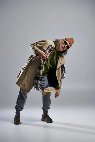 Full length of expressive senior man in dark sunglasses, beanie hat, beige trench coat and plaid scarf looking at camera on grey background, hipster fashion, stylish pose, trendy aging concept — Stock Photo