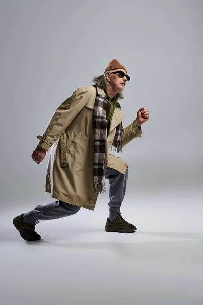 Full length of grey haired man in dark sunglasses and hipster style outfit posing and looking away on grey background, beanie hat, beige trench coat, sneakers, expressive personality — Stock Photo