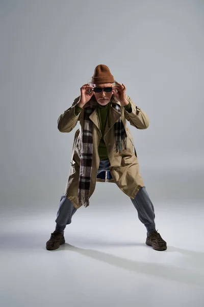 Modisches Senior-Model in stilvoller Pose, gealterter Hipster mit Hut und beigem Trenchcoat, dunkle Sonnenbrille und Blick in die Kamera auf grauem Hintergrund — Stockfoto