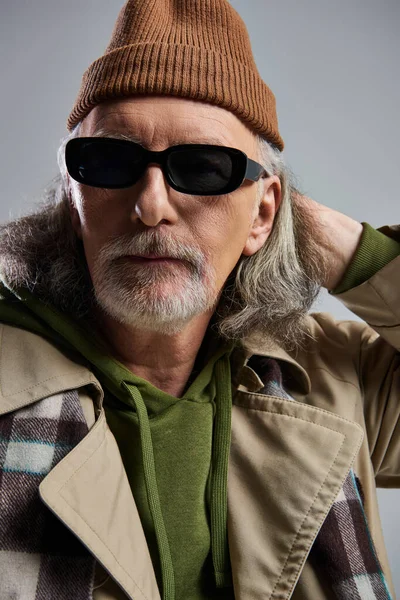 Personnalité expressive, portrait de l'homme âgé de style hipster en bonnet chapeau, lunettes de soleil sombres, trench coat beige et foulard à carreaux regardant la caméra sur fond gris, mode de vie à la mode — Photo de stock