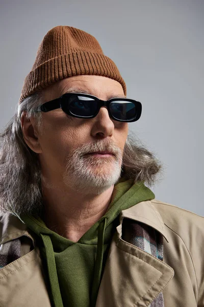 Portrait of confident grey haired and bearded senior man in beanie hat, dark sunglasses and beige trench coat looking away on grey background, hipster style, fashion and age concept — Stock Photo