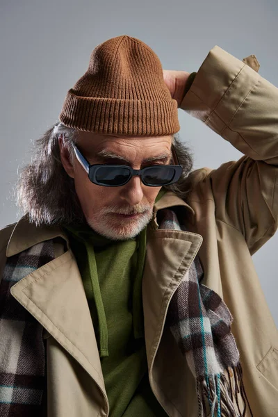 Portrait of senior and bearded man in dark sunglasses, beige trench coat and plaid scarf adjusting beanie hat on grey background, hipster style, fashionable aging concept — Stock Photo