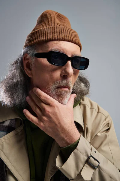 Portrait d'un homme âgé réfléchi au visage découragé, chapeau bonnet, lunettes de soleil sombres et trench coat touchant la barbe sur fond gris, style hipster, tournage de mode — Photo de stock