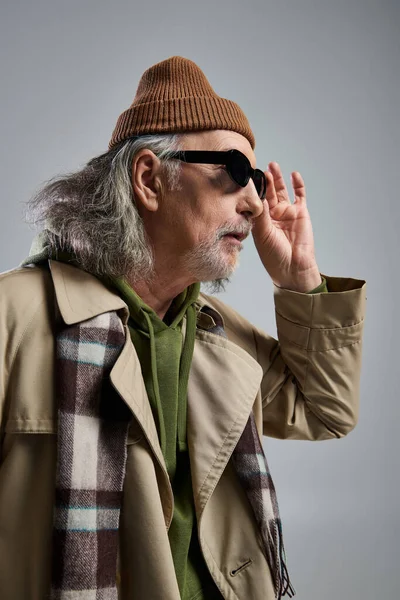 Portrait of amazed elderly man in beanie hat, beige trench coat and plaid scarf adjusting dark sunglasses and looking away on grey background, hipster fashion, stylish and positive aging concept — Stock Photo