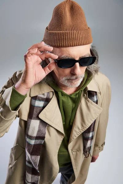 Модный и бородатый мужчина регулирует темные солнцезащитные очки, стоя на сером фоне, хипстерская мода, шляпа шапочка, бежевый плащ, клетчатый шарф, старение с концепцией стиля, модная съемка — стоковое фото