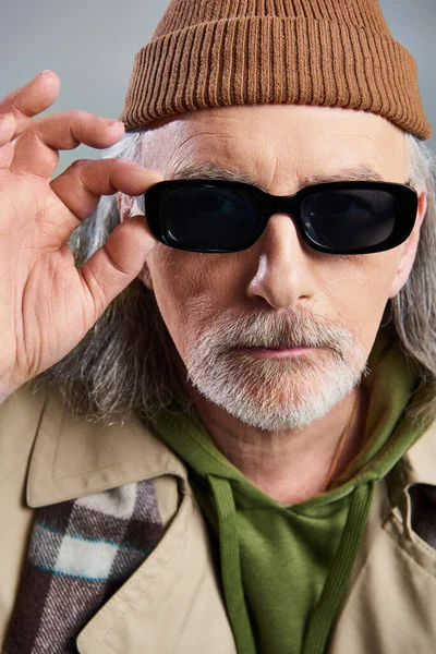 Portrait of senior man with groomed beard, in beanie hat and beige trench coat, touching dark sunglasses and looking at camera on grey background, hipster fashion, individuality, aging with style — Stock Photo