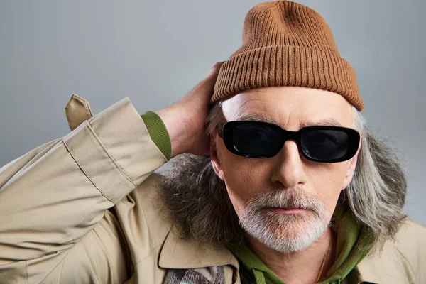Portrait of bearded and hipster style senior man in dark sunglasses, beanie hat and beige trench coat holding hand near head and looking at camera on grey background, trendy aging concept — Stock Photo