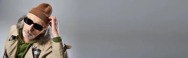 Retrato do modelo masculino sênior em óculos escuros e casaco de trincheira bege ajustando óculos escuros e olhando para a câmera em fundo cinza, homem hipster envelhecido, banner com espaço de cópia — Fotografia de Stock