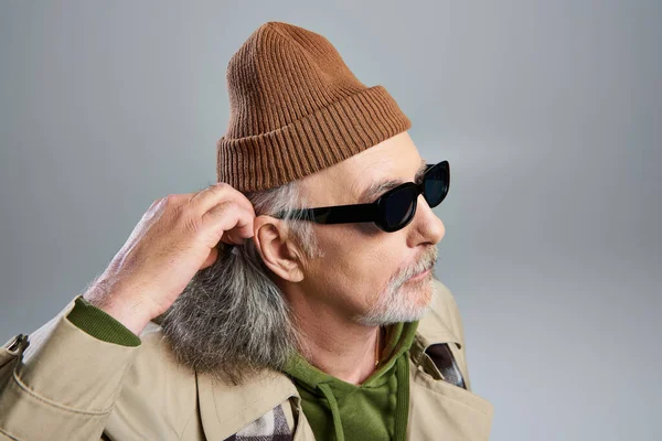 Portrait de vieux et barbu hipster style senior homme en lunettes de soleil sombres, bonnet chapeau et trench coat beige ajuster les cheveux gris et détourner les yeux sur fond gris, concept de vieillissement à la mode — Photo de stock