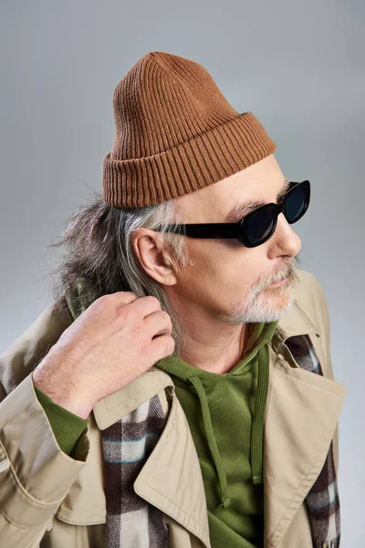 Portrait of aged hipster man in dark sunglasses, beanie hat, green hoodie and beige trench coat looking away while standing on grey background, aging with style concept, fashion shoot — Stock Photo