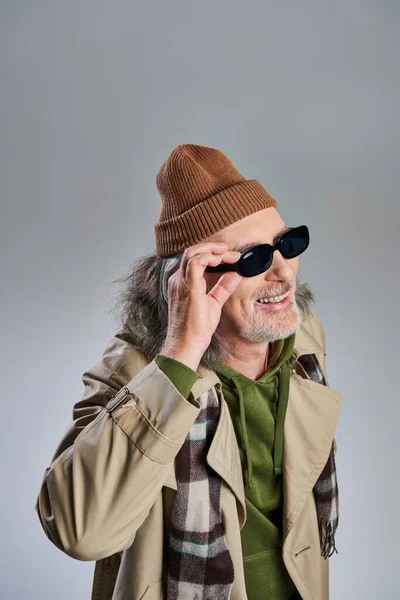 Retrato de hombre barbudo de estilo hipster sénior y alegre en gorro sombrero y gabardina beige ajustando gafas de sol oscuras, riendo y mirando hacia otro lado sobre fondo gris - foto de stock