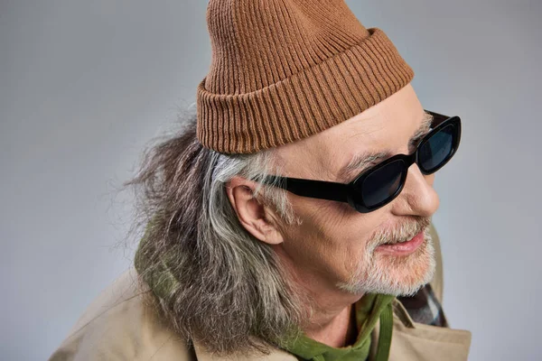 Retrato de hombre mayor de pelo gris y alegre en traje de estilo hipster de moda, gorro sombrero y gafas de sol oscuras sonriendo y mirando hacia otro lado sobre fondo gris, concepto de envejecimiento feliz - foto de stock