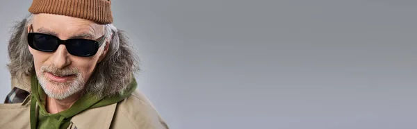 Individualidade expressiva, retrato de homem sênior sorridente em chapéu de gorro, óculos escuros elegantes e casaco de trincheira bege sorrindo para a câmera em fundo cinza, envelhecimento positivo, banner com espaço de cópia — Fotografia de Stock