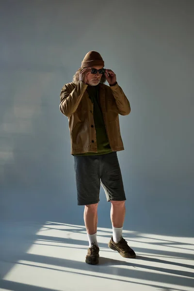 Повна довжина модного старшого чоловіка, що регулює темні сонцезахисні окуляри, стоячи в капелюсі, куртці та шортах на сірому фоні з освітленням, хіпстерська мода, позитивна та модна концепція старіння — стокове фото