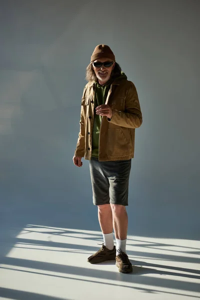 Full length of cheerful senior man in beanie hat, dark sunglasses, jacket and shorts smiling at camera on grey background with lighting, hipster style, happy and fashionable aging concept — Stock Photo