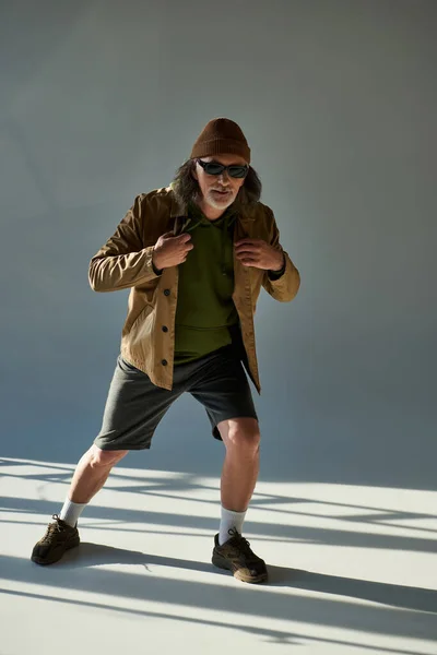 Älterer Mann im Hipster-Look mit Hut, dunkler Sonnenbrille, Jacke und kurzen Hosen, die auf grauem Hintergrund mit Beleuchtung in die Kamera schauen, stilvolle Pose, trendiges alterndes Konzept — Stockfoto