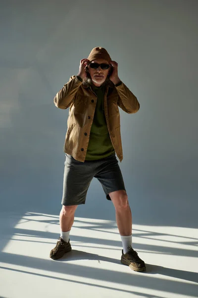 Expressive personality, hipster fashion, full length of elderly man in dark sunglasses, jacket and shorts adjusting beanie hat and looking at camera on grey background with lighting — Stock Photo