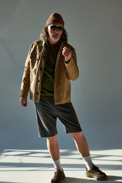 Vollbärtiger Mann mit Hut, dunkler Sonnenbrille, Jacke und kurzen Hosen, der in die Kamera blickt, während er auf grauem Hintergrund mit Beleuchtung steht, Hipster-Mode, trendiges alterndes Konzept — Stockfoto