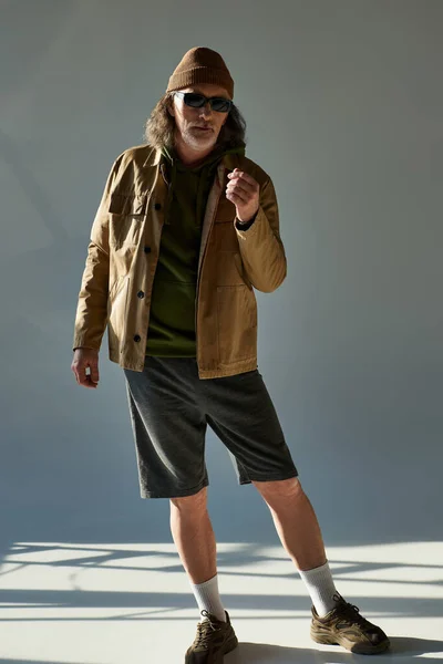 Longitud completa del hombre barbudo envejecido en sombrero de gorro, gafas de sol oscuras, chaqueta y pantalones cortos mirando a la cámara mientras está de pie sobre fondo gris con iluminación, moda hipster, concepto de envejecimiento de moda - foto de stock