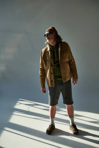 Vollbärtiger älterer Mann mit dunkler Sonnenbrille, Hut, Jacke und kurzen Hosen auf grauem Hintergrund mit Beleuchtung, Hipster-Trend, ausdrucksstarke Persönlichkeit — Stockfoto