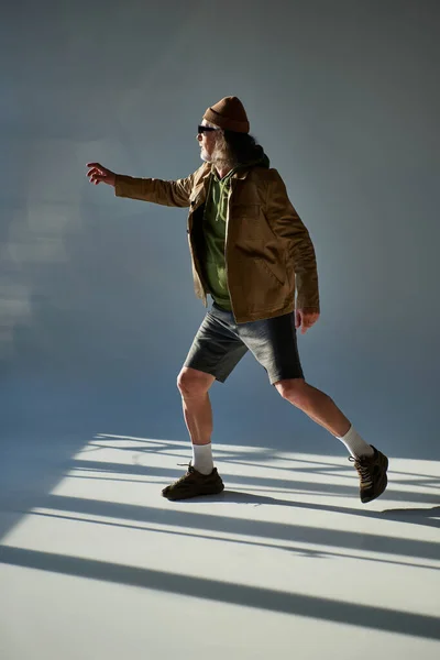 Full length of senior man in fashionable casual clothes, dark sunglasses, beanie hat, jacket and shorts pointing with hand and looking away on grey background with lighting, aging with style concept — Stock Photo