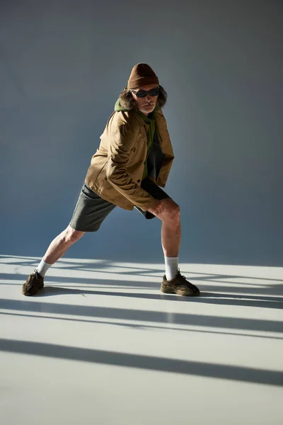 Expressive and trendy personality, full length of senior man in dark sunglasses, beanie hat, jacket and shorts looking at camera while posing on grey background with lighting — Stock Photo