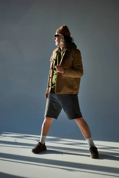 Älterer, grauhaariger Mann mit Mütze, dunkler Sonnenbrille, Jacke und kurzen Hosen, der vor grauem Hintergrund mit Beleuchtung im Hipster-Stil steht und wegschaut. — Stockfoto