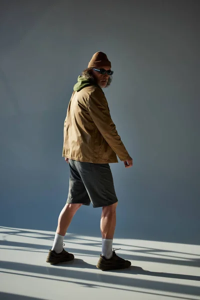 Полная длина хипстерский стиль старший человек в шляпе шапочка, темные солнцезащитные очки, куртка и шорты глядя в сторону, стоя на сером фоне с освещением, модная концепция старения — стоковое фото